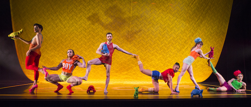 Dancers in colorful athletic wear arrange themselves downstage all connecting to one another in some way. A man wears pink high heals. Another squats turned out will a dancer rests his foot on his thigh.
